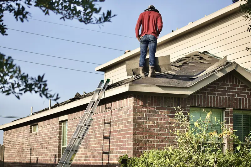 Roofer standing on a damaged shingle roof of a brick house, inspecting the problem area with a ladder against the wall